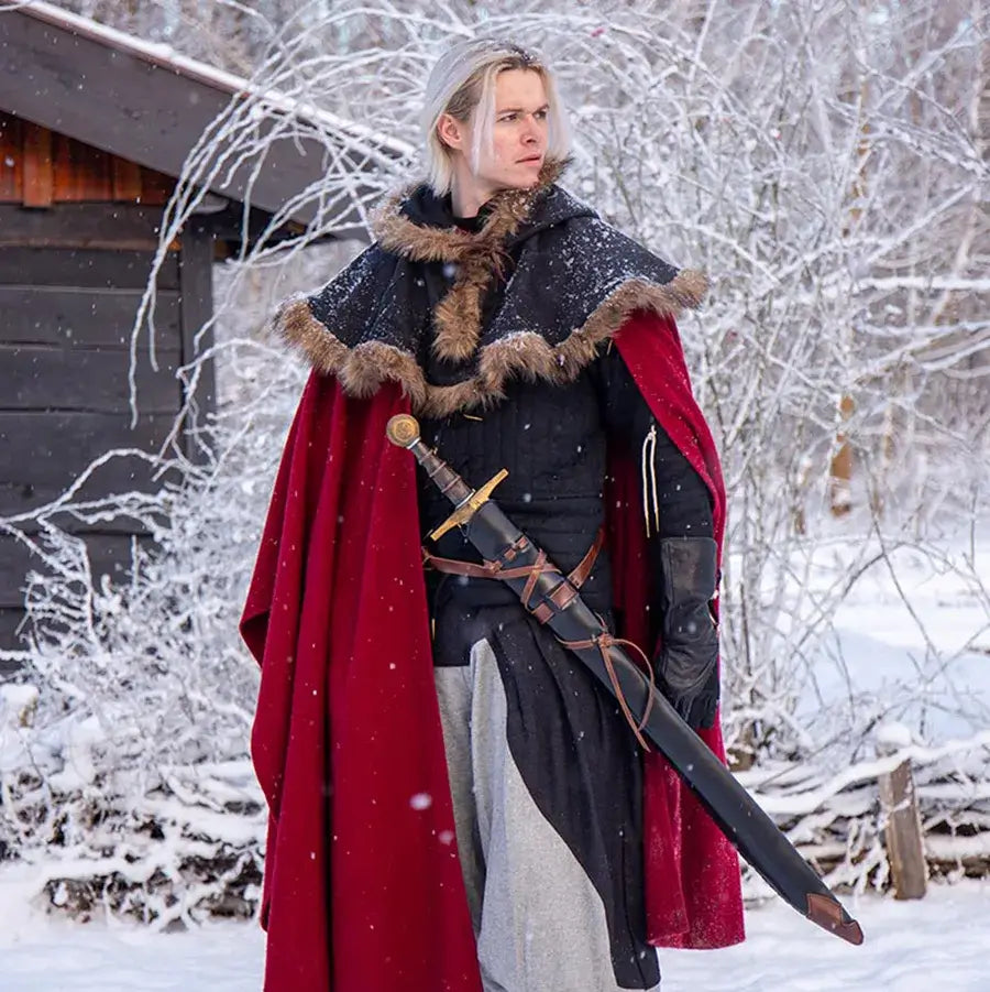 A warrior in a red cape stands in the snow, showcasing elegant Scabbard Solutions with a sword by their side.