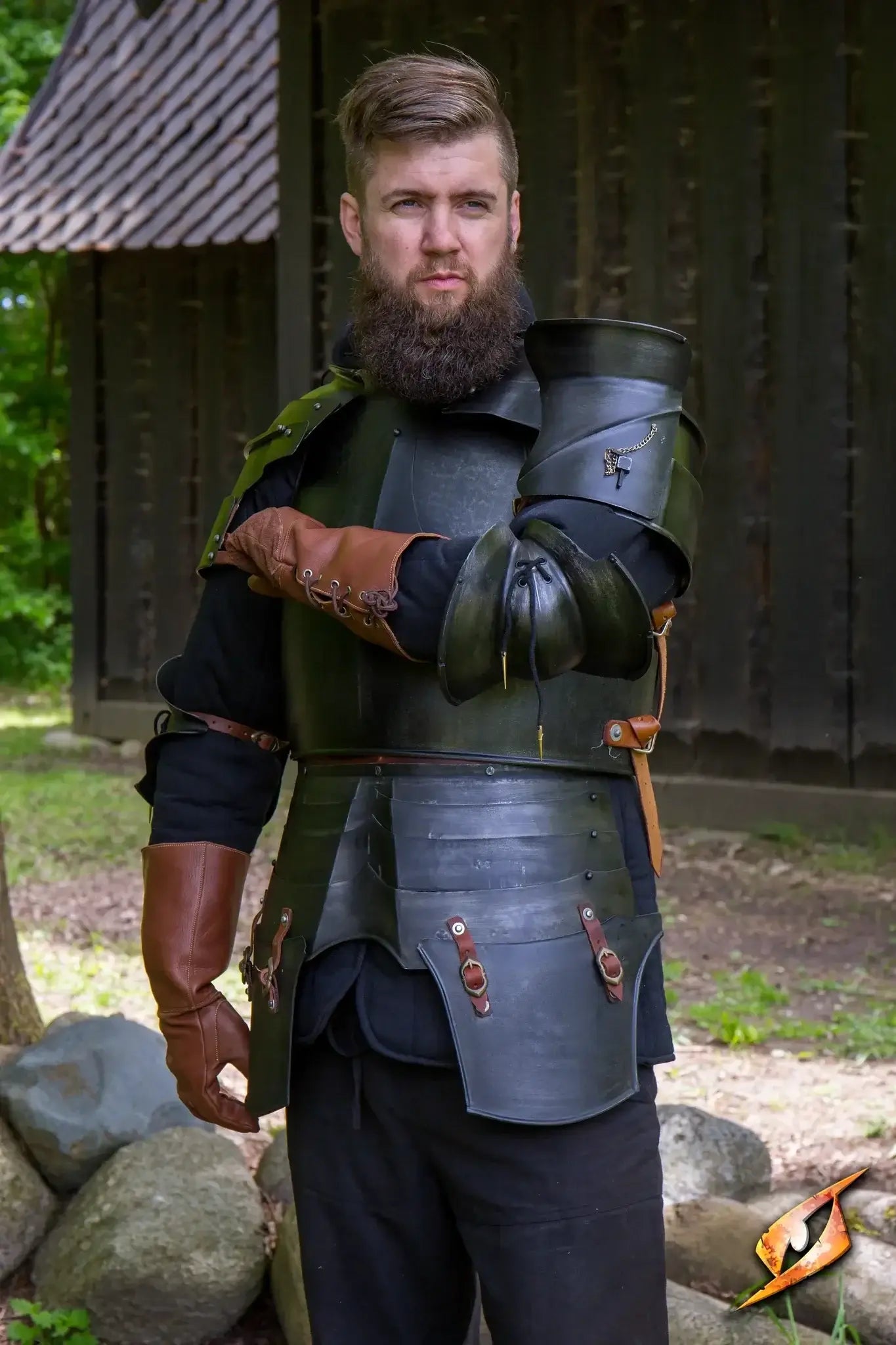 Man in black plate armor standing with a wooden house as backdrop