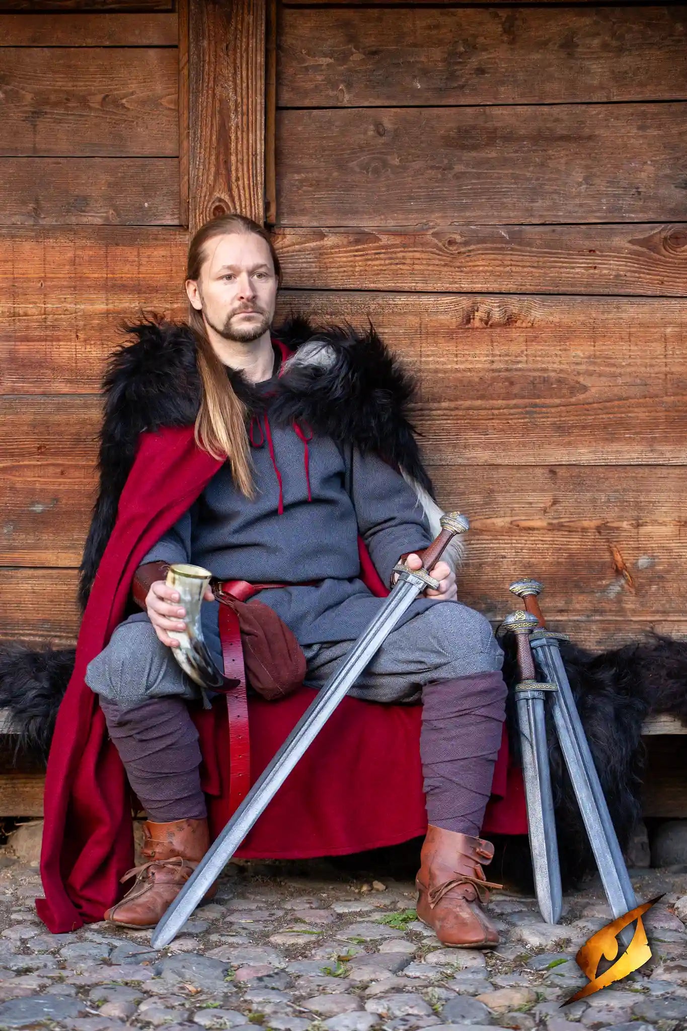 A model wearing Viking Clothes, showcasing Scandinavian style and durability, seated with swords and a cup.