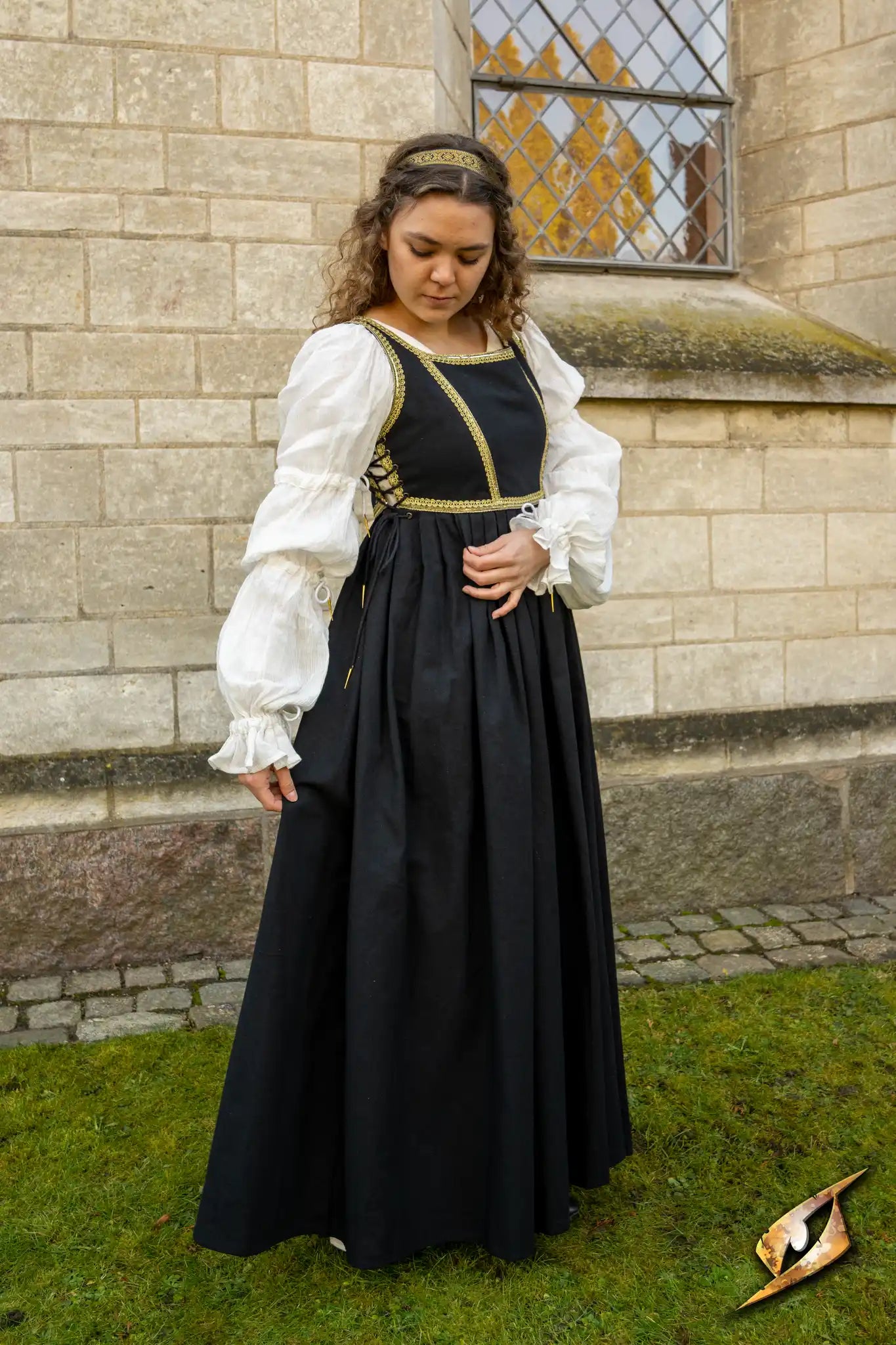 Model wearing Dress Lucrezia, a black gown with golden trimmings and white sleeves, standing by a stone wall.
