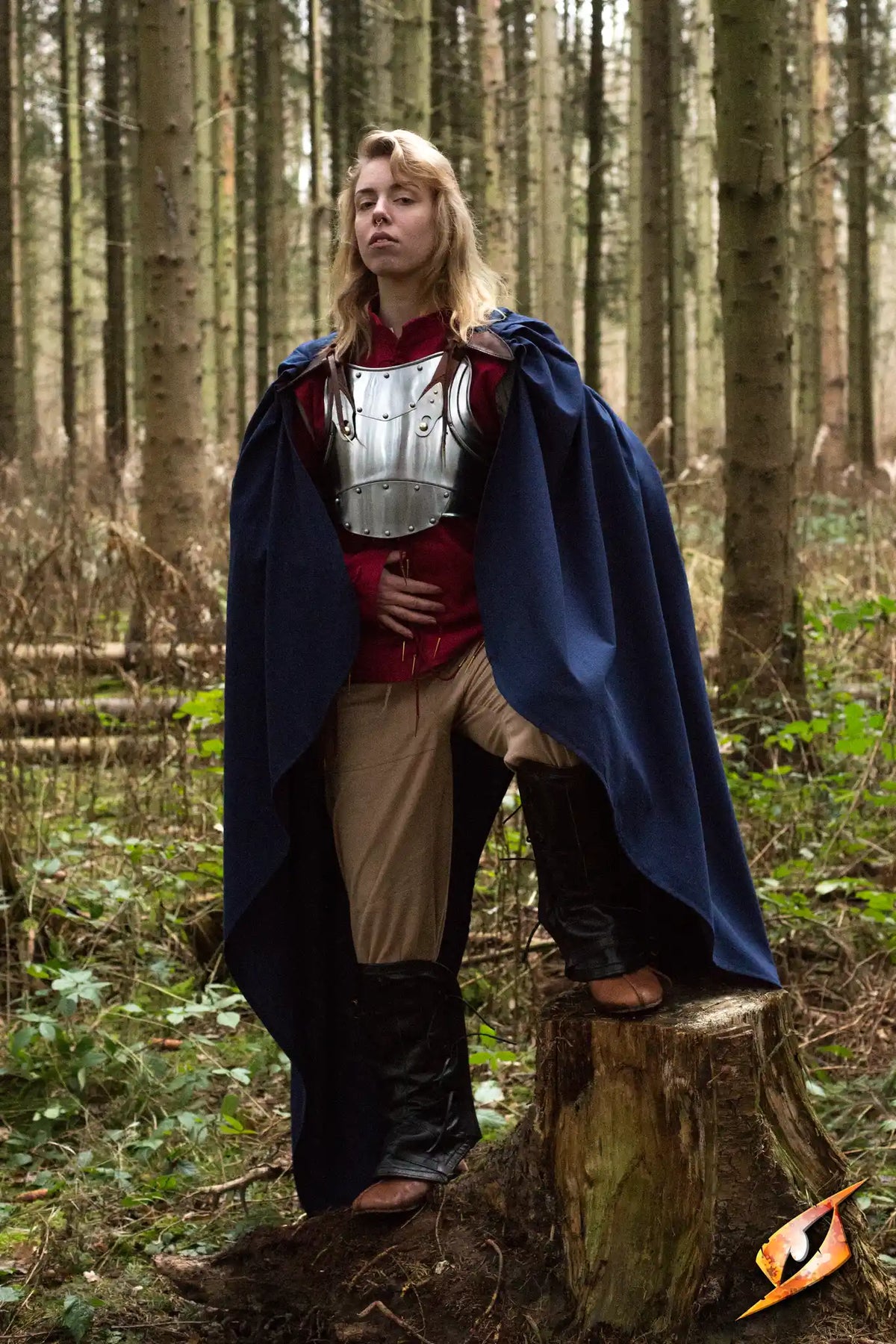 A model wearing the Cape Regulus, a medium-length blue cape, in a forest setting, showcasing its elegant design and protection.