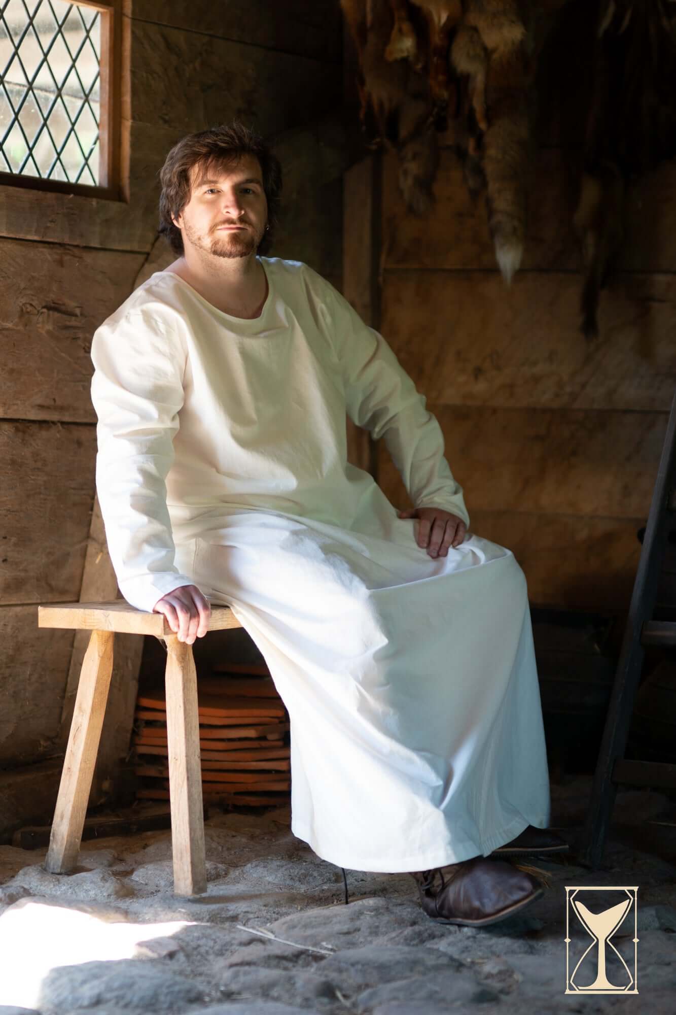 A man wearing the Undertunic Modesto, seated on a wooden stool in a rustic setting, showcasing its historical design.