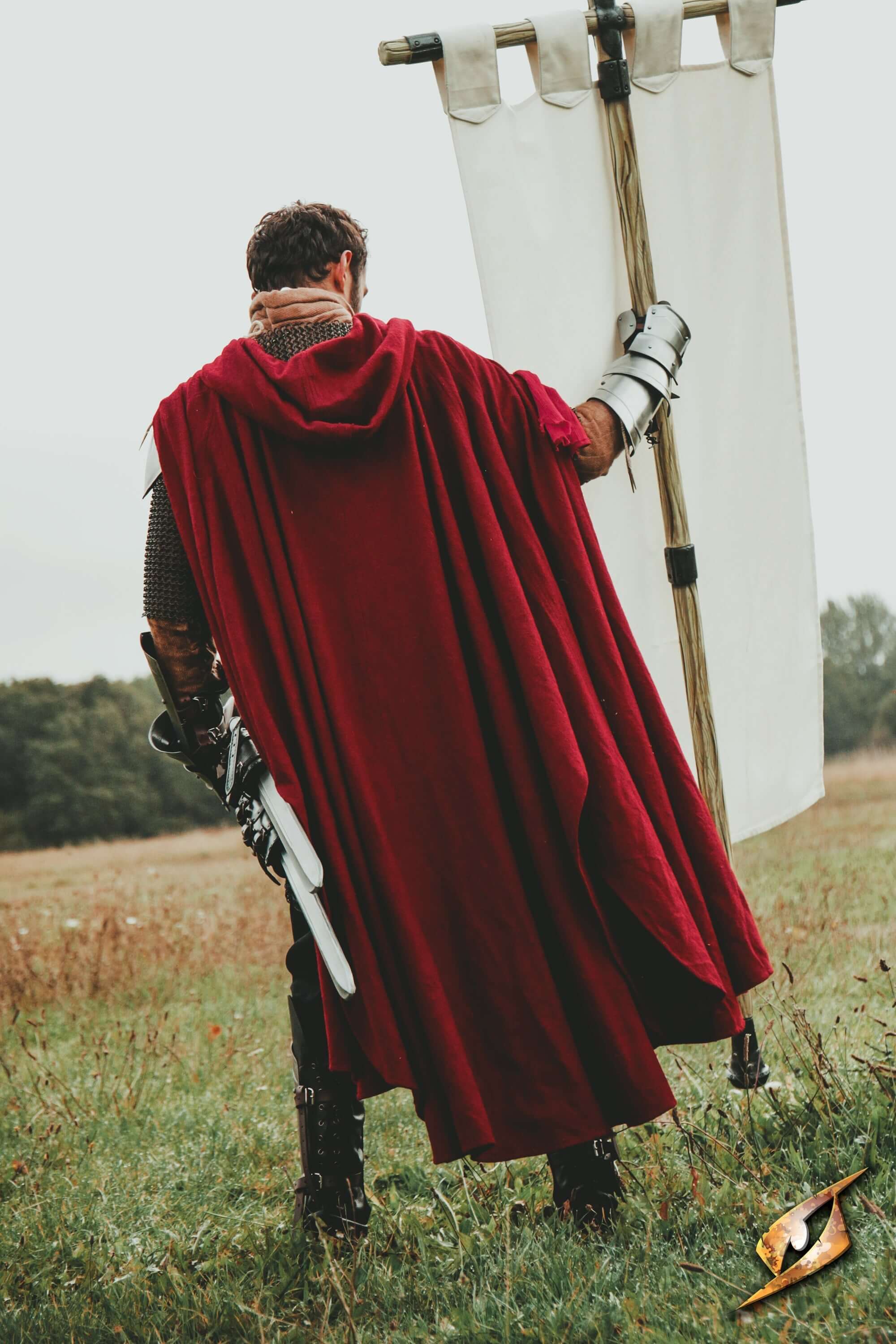 Man in a red cape standing in a field holding a banner, showcasing Capes & Cloaks in an outdoor setting.