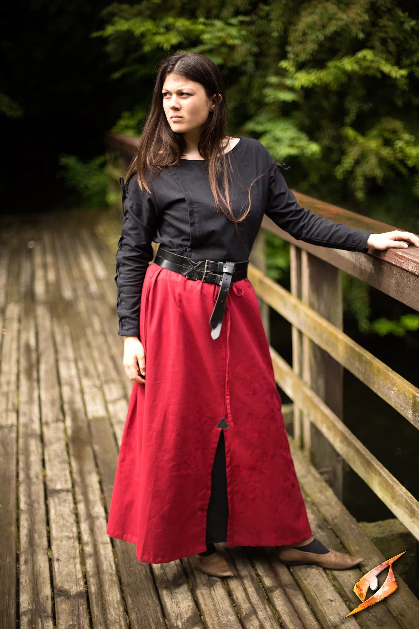 Model wearing a red Battle Skirt with a black top, standing on a wooden bridge surrounded by greenery.