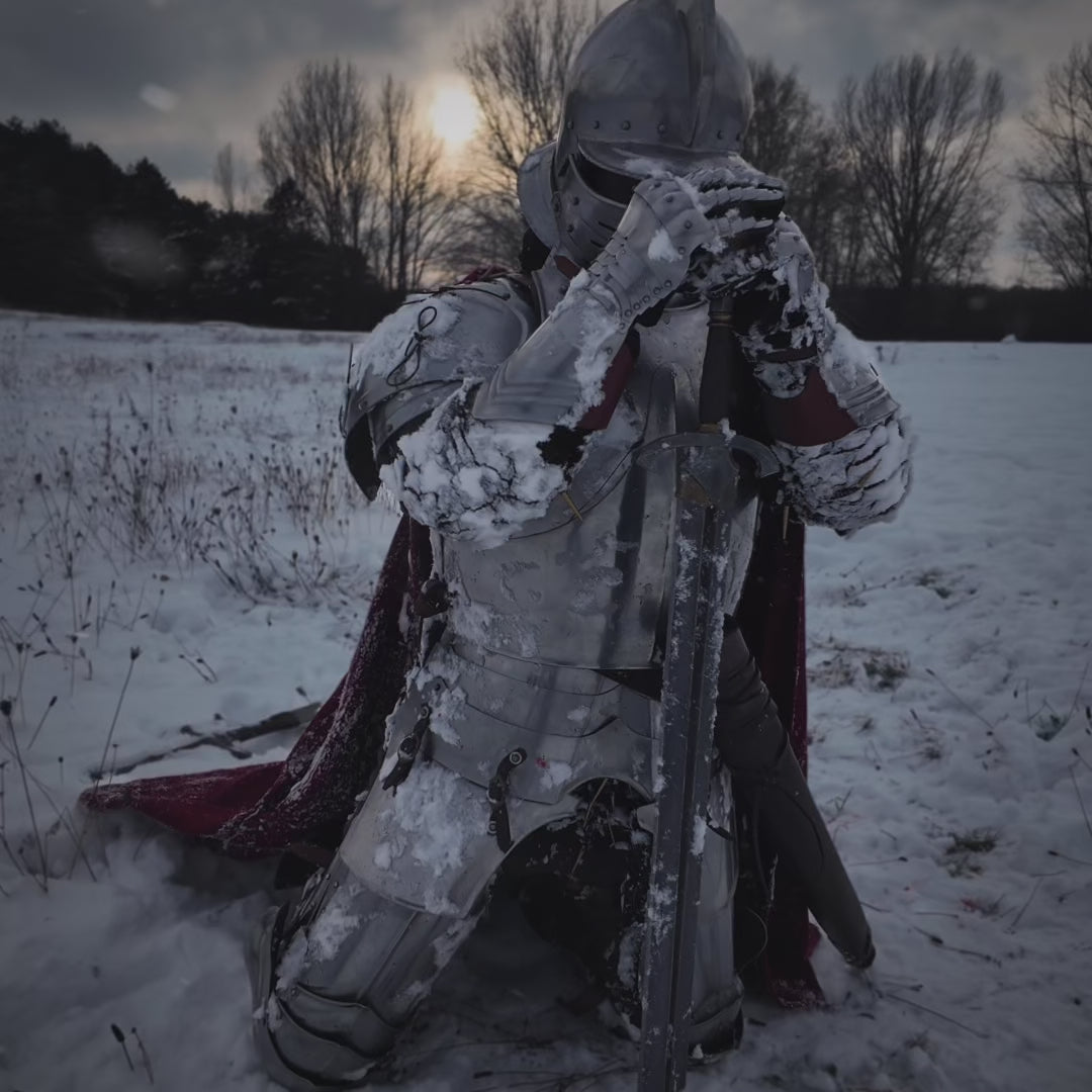Knight in steel plate armor and helmet in a snowy winter landscape, holding a sword and wearing medieval gauntlets and a red wool cloak. Filmed outdoors in cold weather. 